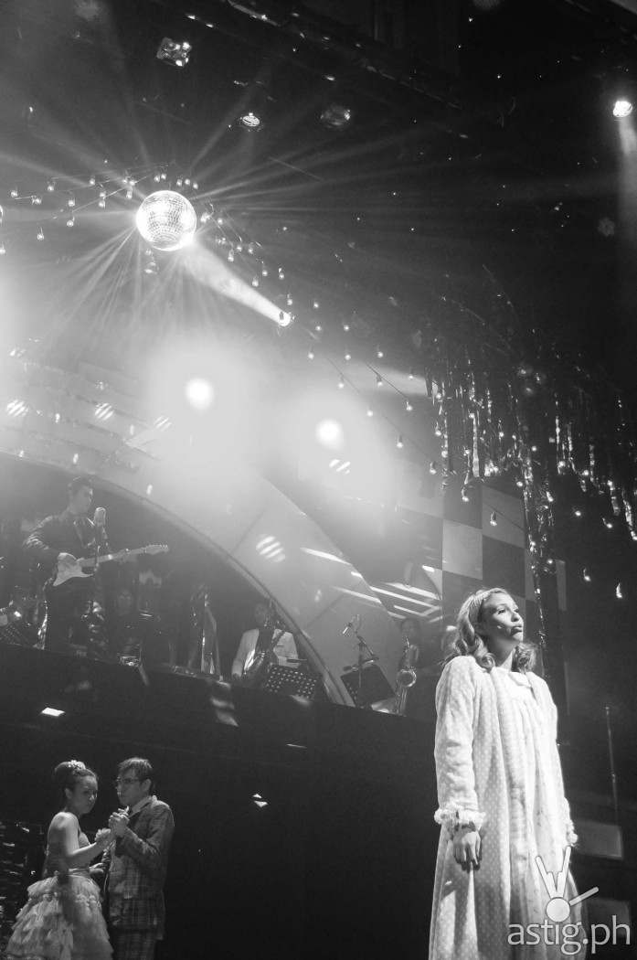 Sandy Dumbrowski (Tippy Dos Santos) sings while a glitterbal illuminates the stage and couples dance in the background